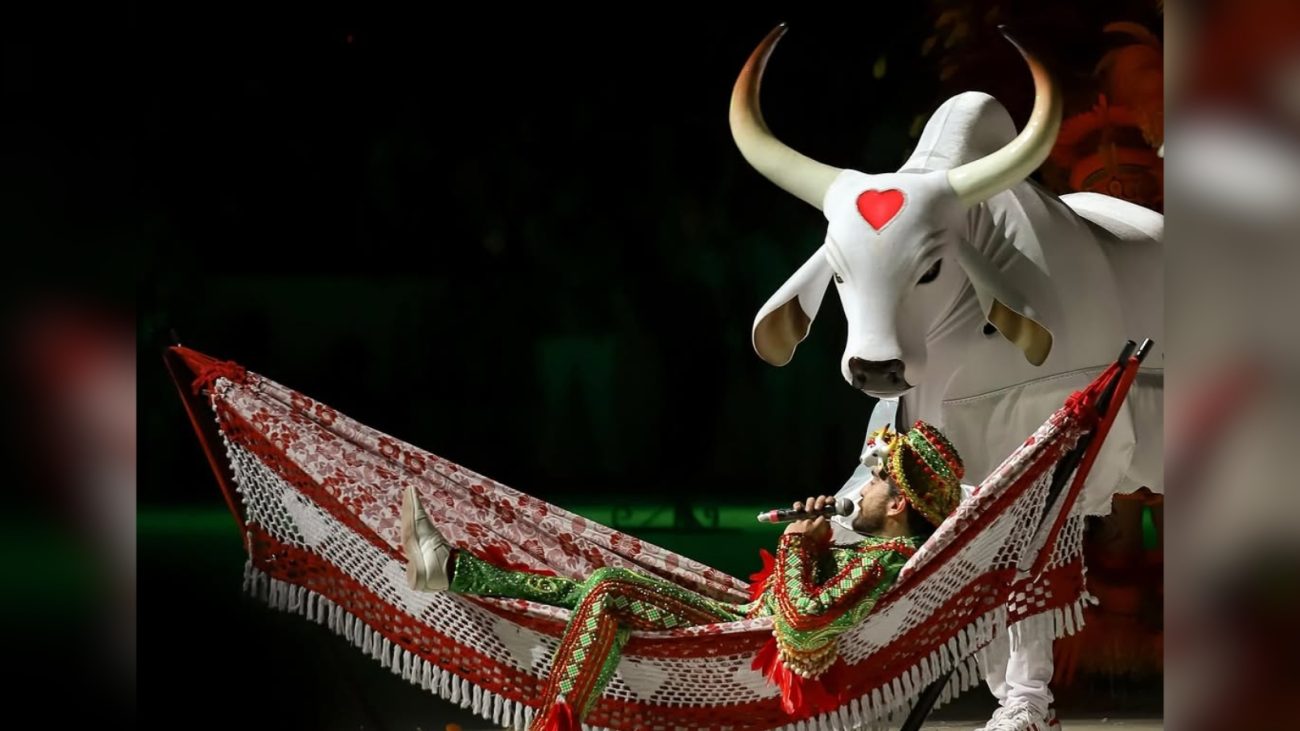 Cena de uma apresentação cultural com uma pessoa vestida com roupas indígenas e uma máscara de touro gigante ao fundo, simbolizando a cultura brasileira.